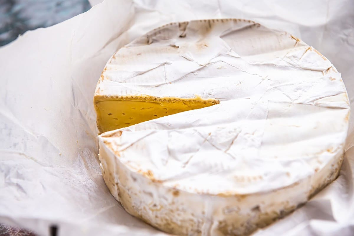 French soft brie cheese round wheel in paper package, dairy double cream product on kitchen table macro closeup with cut slice