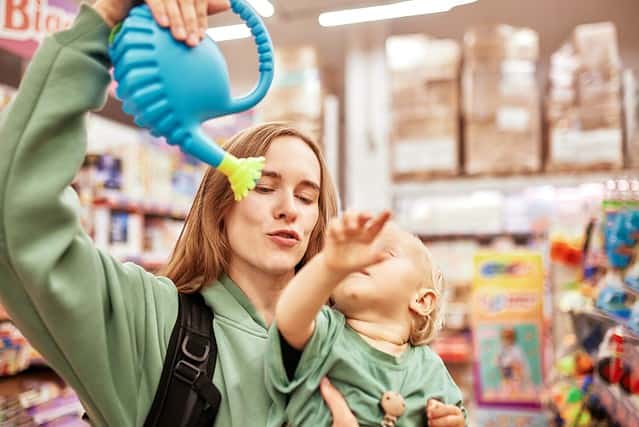 Happy young mother with cute baby in her hands walking around toy store in shopping mall. Mom and little blonde daughter choosing toys and having fun. Funny family weekend, happy childhood.