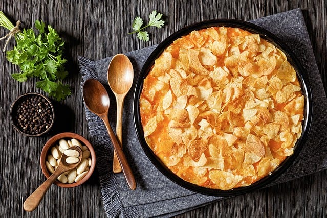 creamy cheesy chicken almond casserole topped with potato chips in baking dish on dark wooden table with ingredients, american recipe, horizontal view from above, flat lay