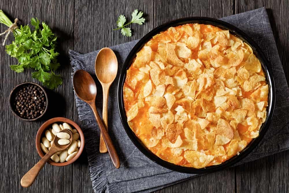 creamy cheesy chicken almond casserole topped with potato chips in baking dish on dark wooden table with ingredients, american recipe, horizontal view from above, flat lay