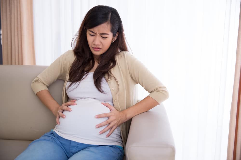 A portrait of an Asian pregnant woman has stomachache sitting on her couch