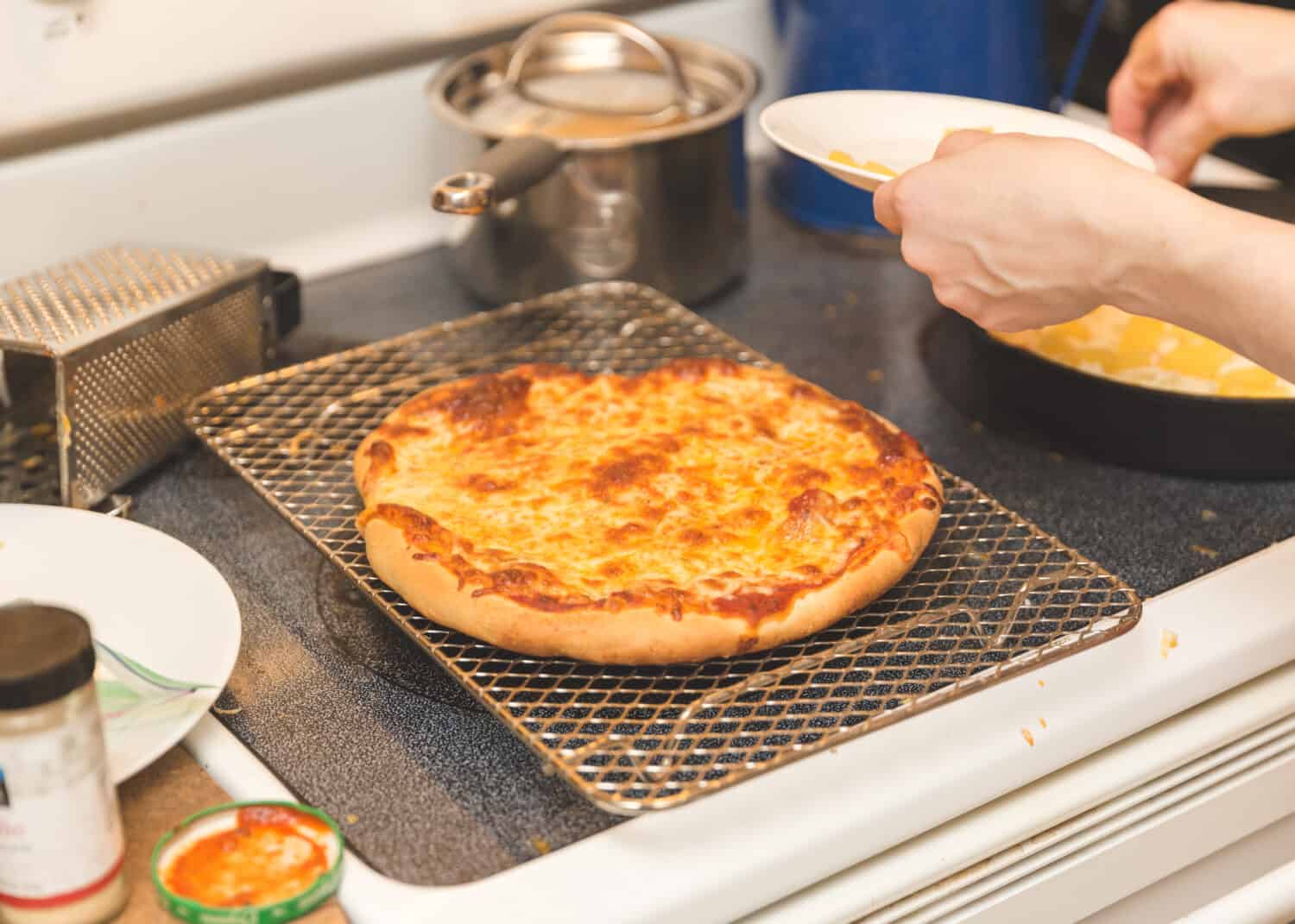 Homemade cheese pizza on baking rack on stovetop. Person preparing food nearby.