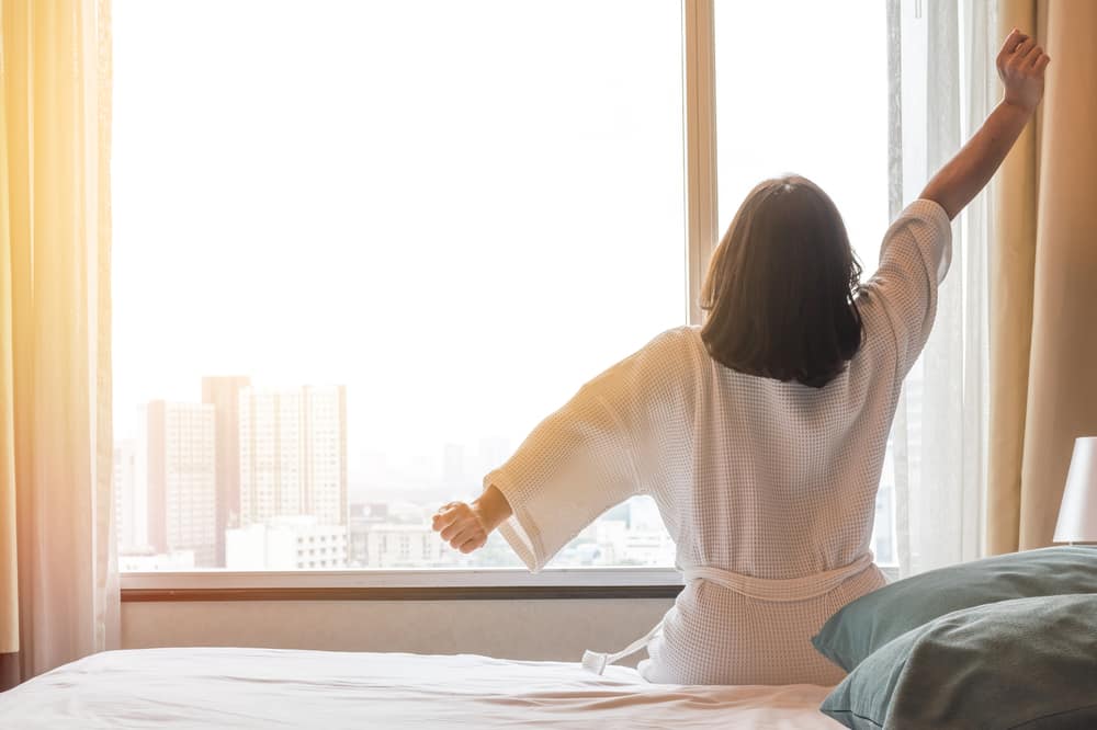 Easy lifestyle Asian woman waking up from good sleep in weekend morning taking some rest, relaxing in comfort bedroom at hotel window, having happy lazy day enjoying work-life quality balance concept