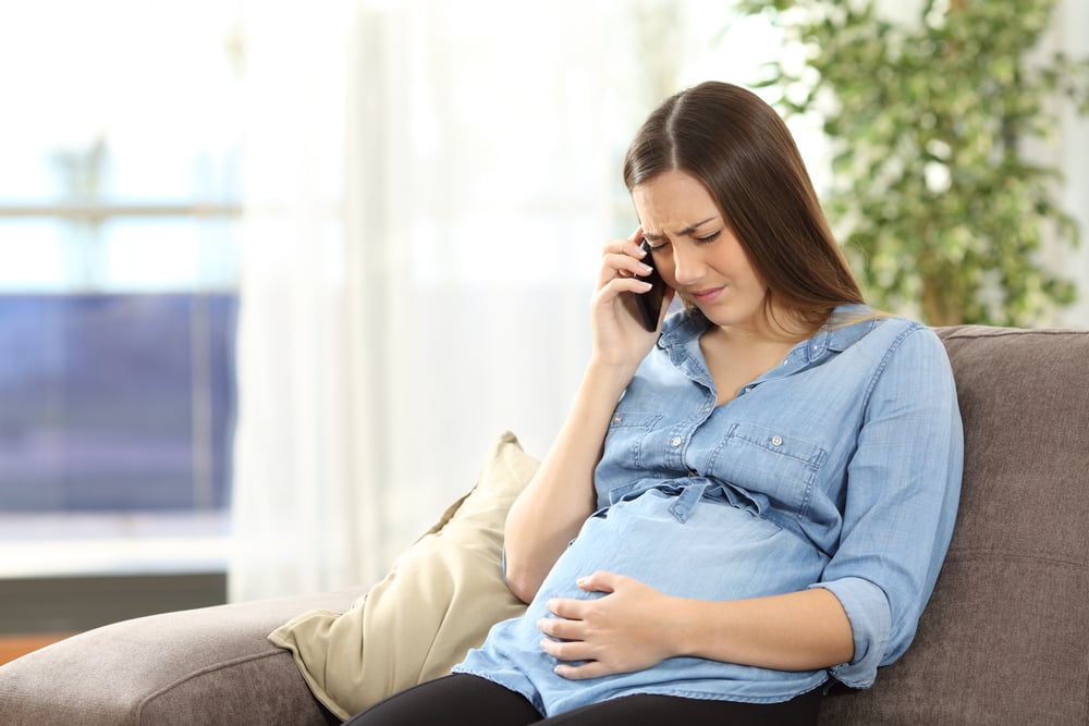 Young pregnant woman suffering belly ache and calling on the phone sitting on a couch in the living room in a house interior