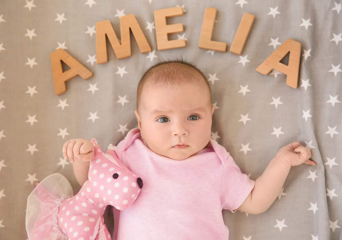 Cute baby lying on bed with toy and word AMELIA composed of wooden letters. Choosing name concept