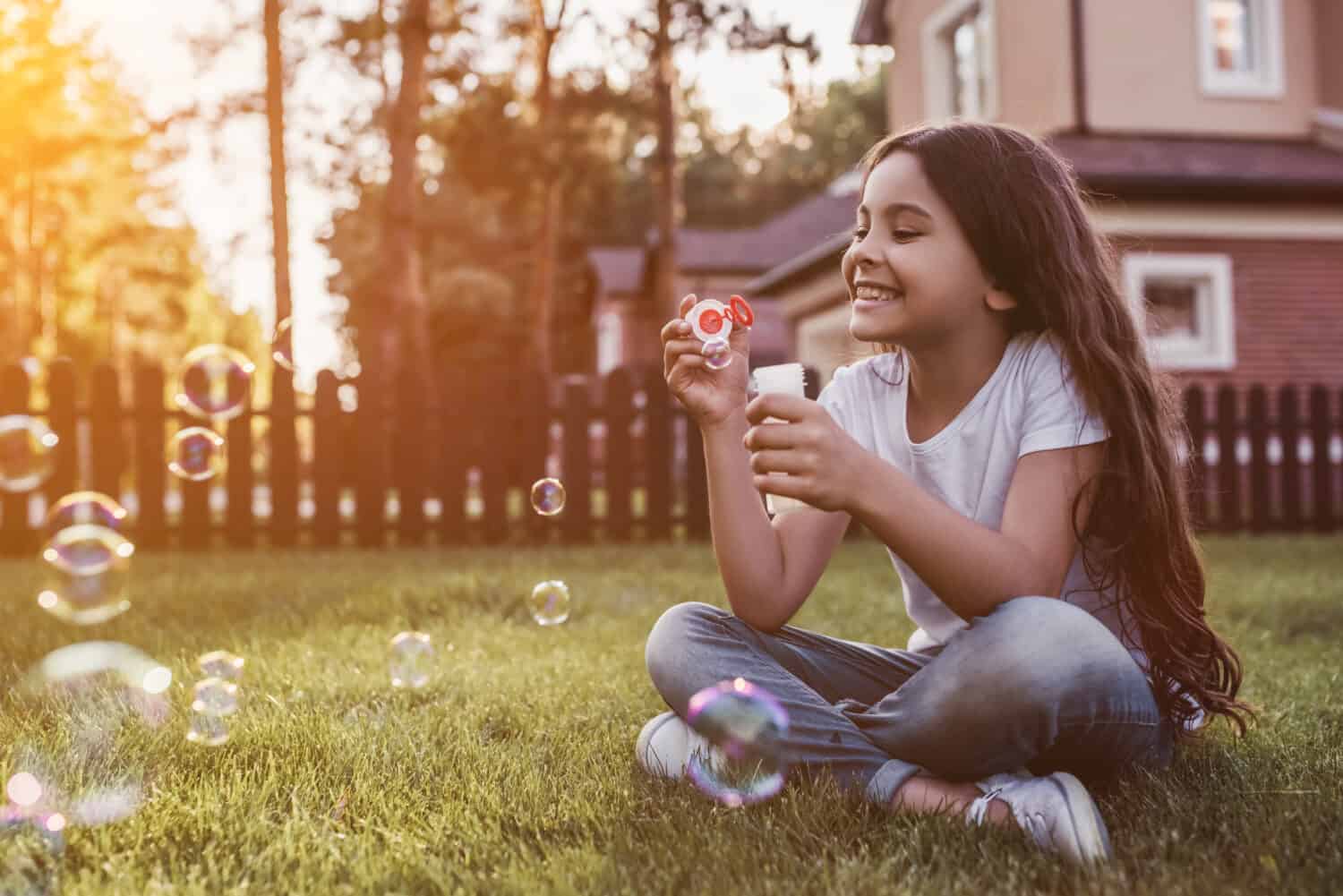 Pretty little girl is sitting on the green grass on the backyard and blowing soap bubbles. Having fun and smiling on the sunset.