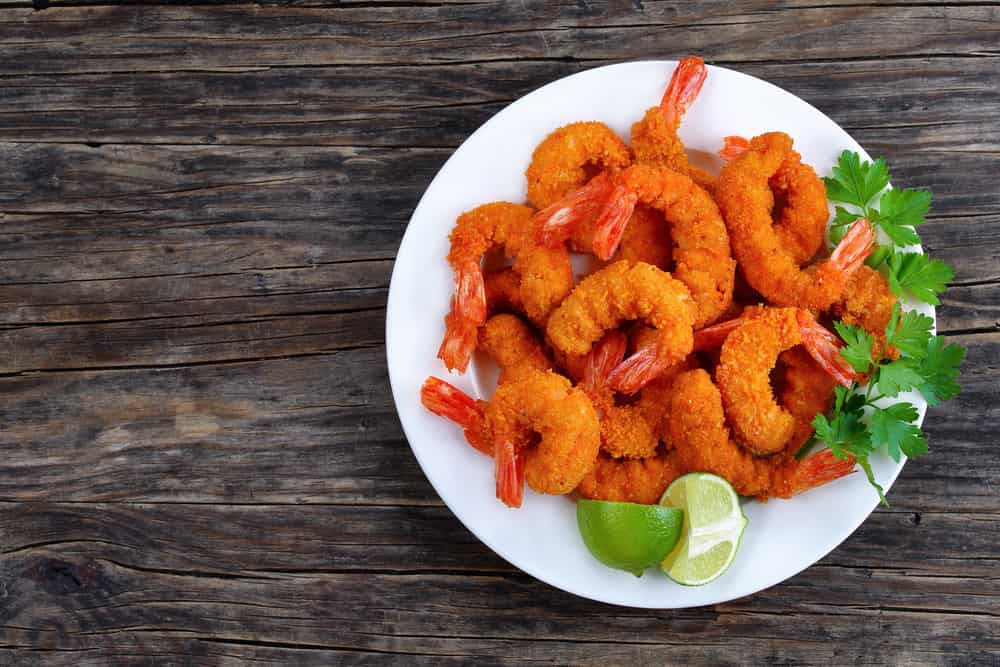 delicious deep fried breaded shrimps served with lime wedges and coriander leaves on white plate on wooden table, copy space for text, view from above