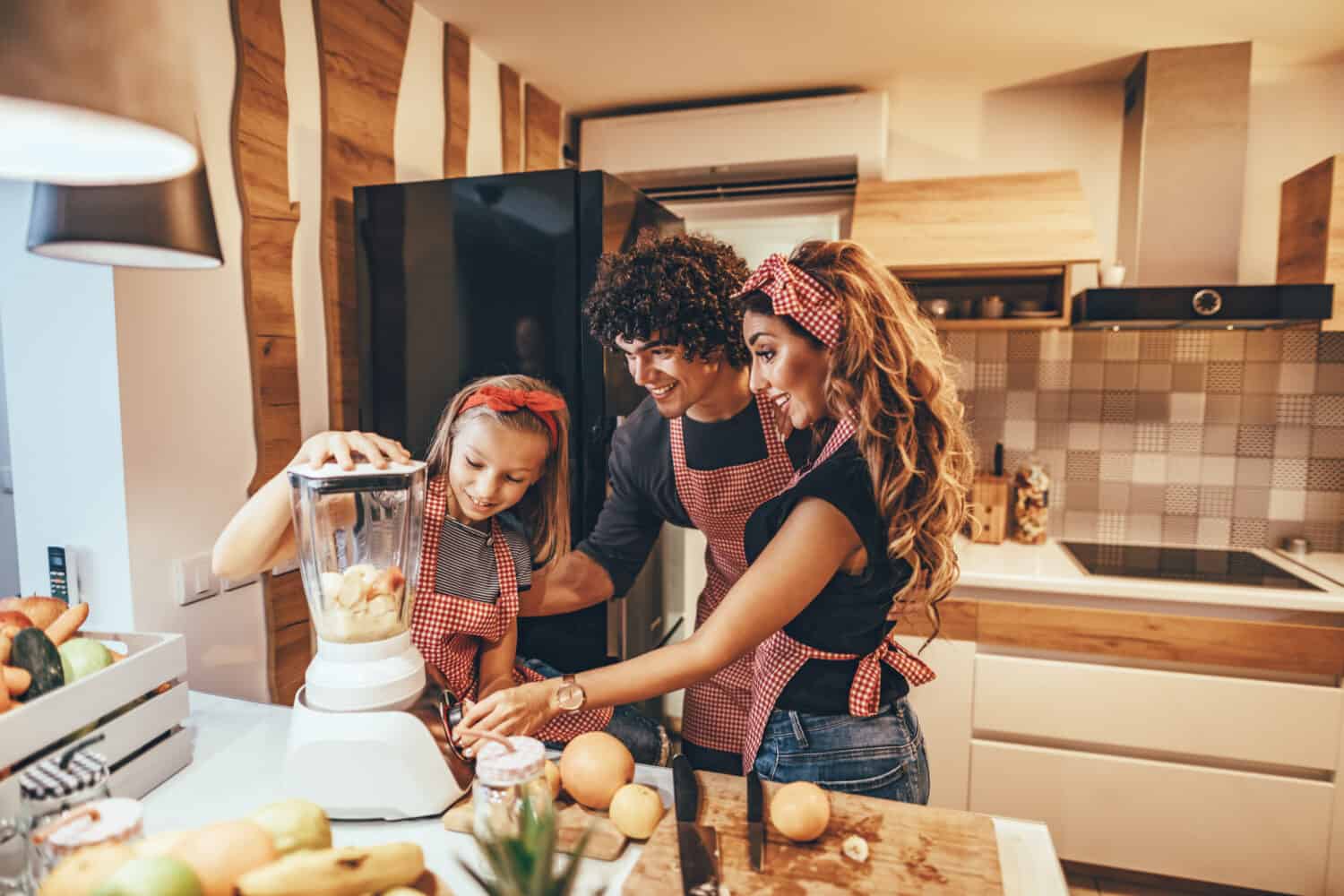 Happy young family is preparing healthy meal in the kitchen. Mother, father and daughter are putting slices of fruit in the blender and making fruit smoothie.