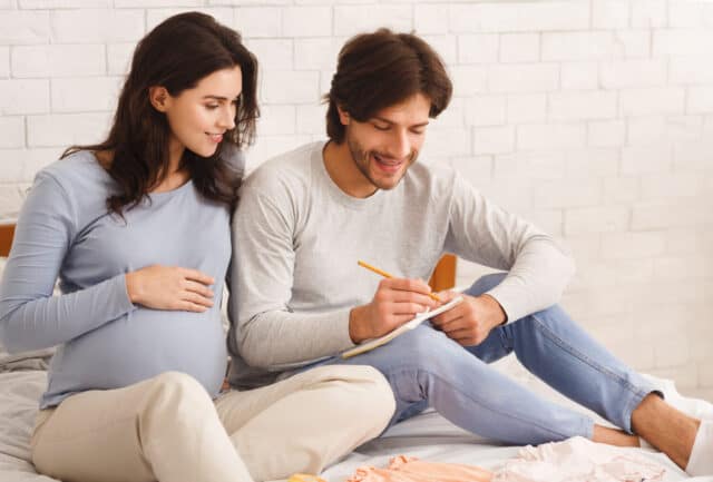 Choosing Baby Name. Young pregnant couple naming their future child, writing options to notebook while sitting on bed at home