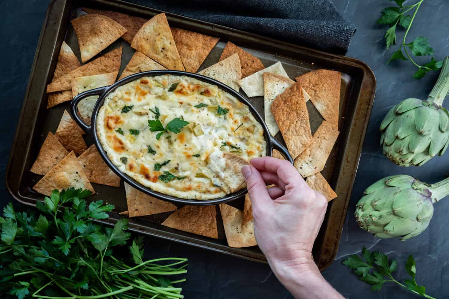 Top down view of a hand dipping a pita triangle into a warm artichoke dip.