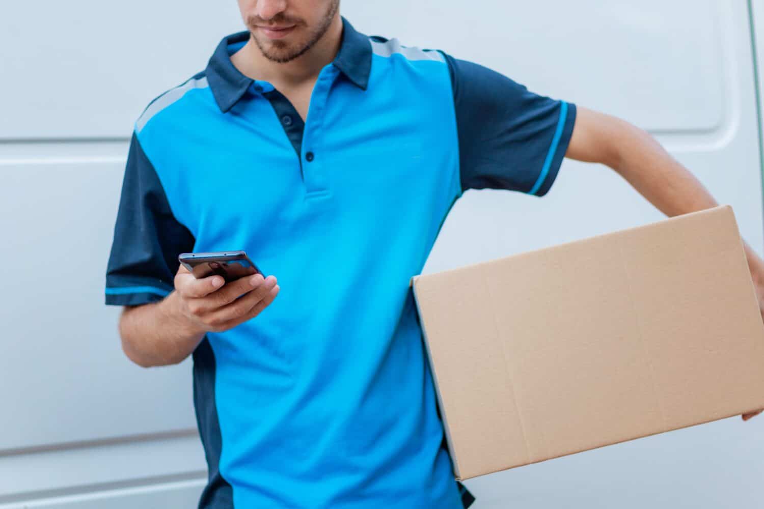 young, slim, handsome, white delivery boy delivering a box and in front of van. Smiling, relaxed. Using the mobile