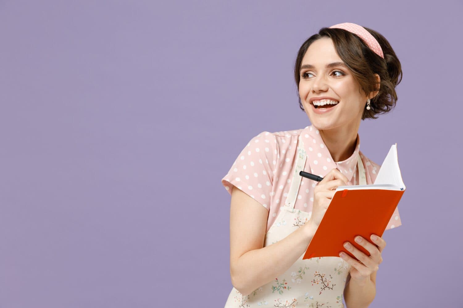 Young amazed housewife housekeeper chef cook baker woman wearing pink apron write down recipe in cook book notebook look aside isolated on pastel violet background studio Cooking food process concept