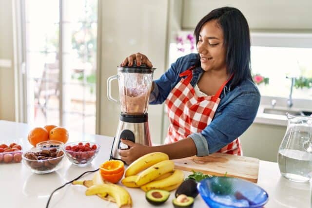 Hispanic brunette woman preparing fruit smoothie with blender at the kitchen