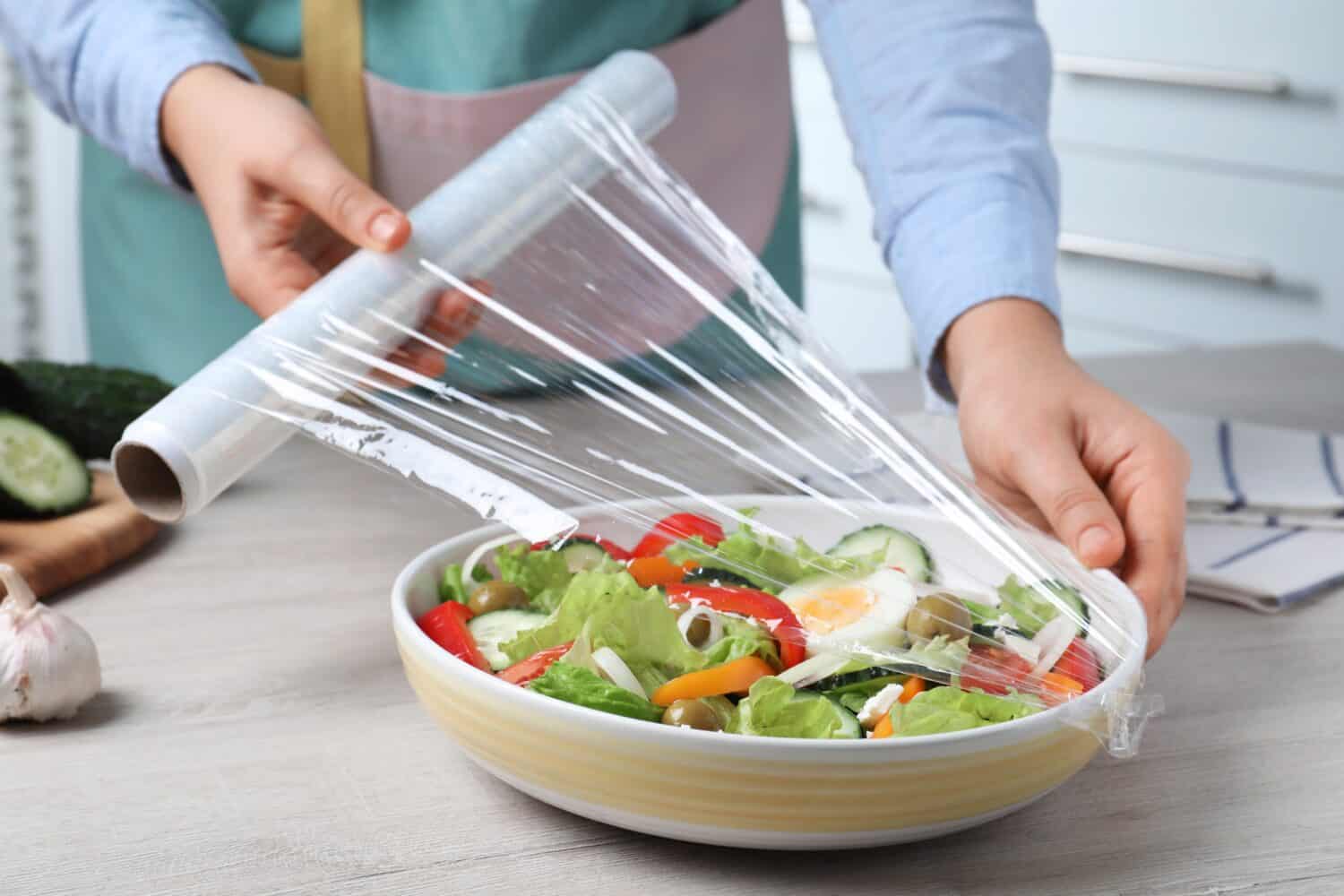 Woman putting plastic food wrap over bowl of fresh salad at wooden table indoors, closeup