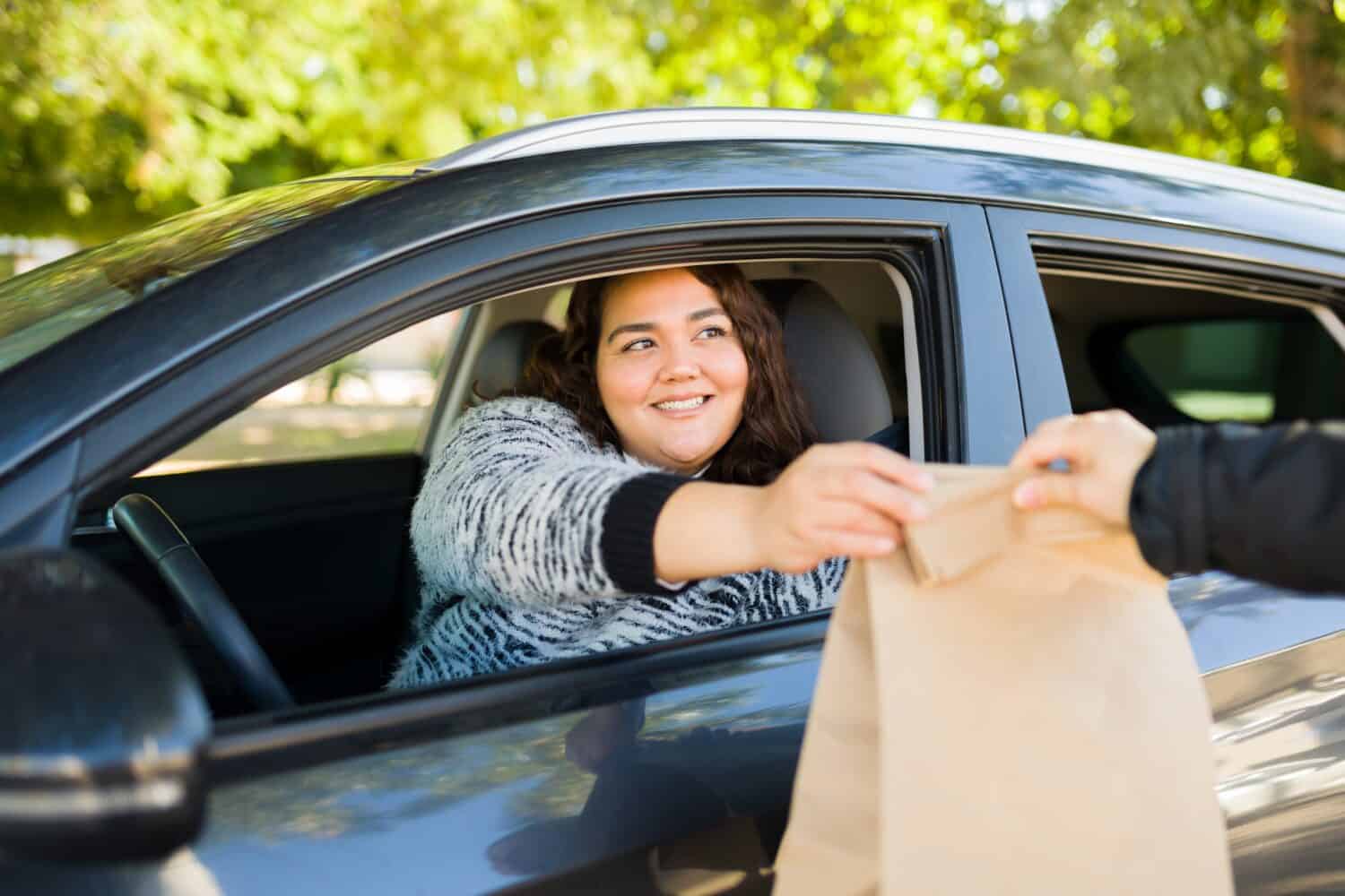 Cheerful obese woman smiling while buying fast food at a drive thru while driving her car 