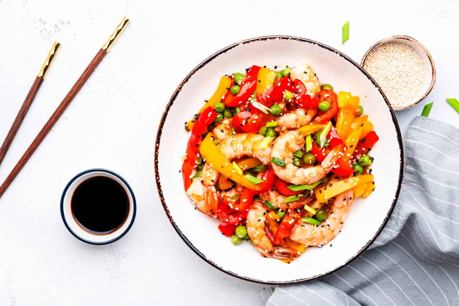 Stir fry with shrimps, red and yellow paprika, green pea, chives and sesame seeds in white bowl Asian cuisine dish. Light stone kitchen table background, top view