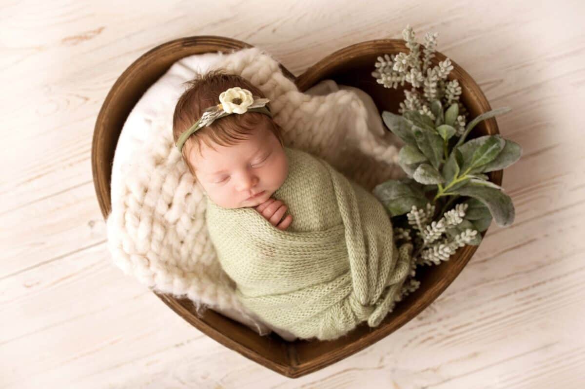 A cute newborn baby in a green pistachio winding and a white bandage with a flower on his head sleeps sweetly. Wooden basket in the shape of heart. Professional macro photo against light wooden floor.