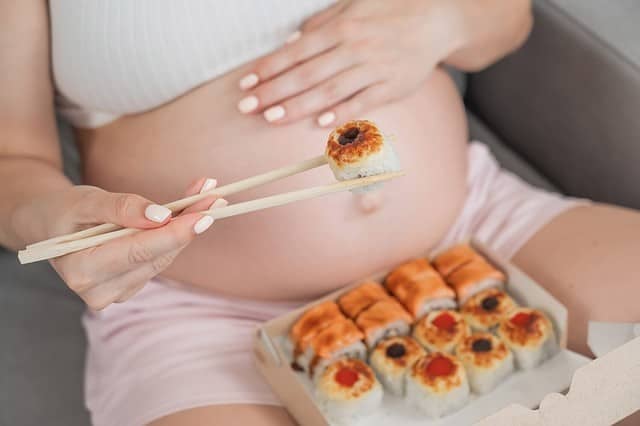 A pregnant woman sits on the sofa and eats rolls from a box. Food delivery. Close up of belly.
