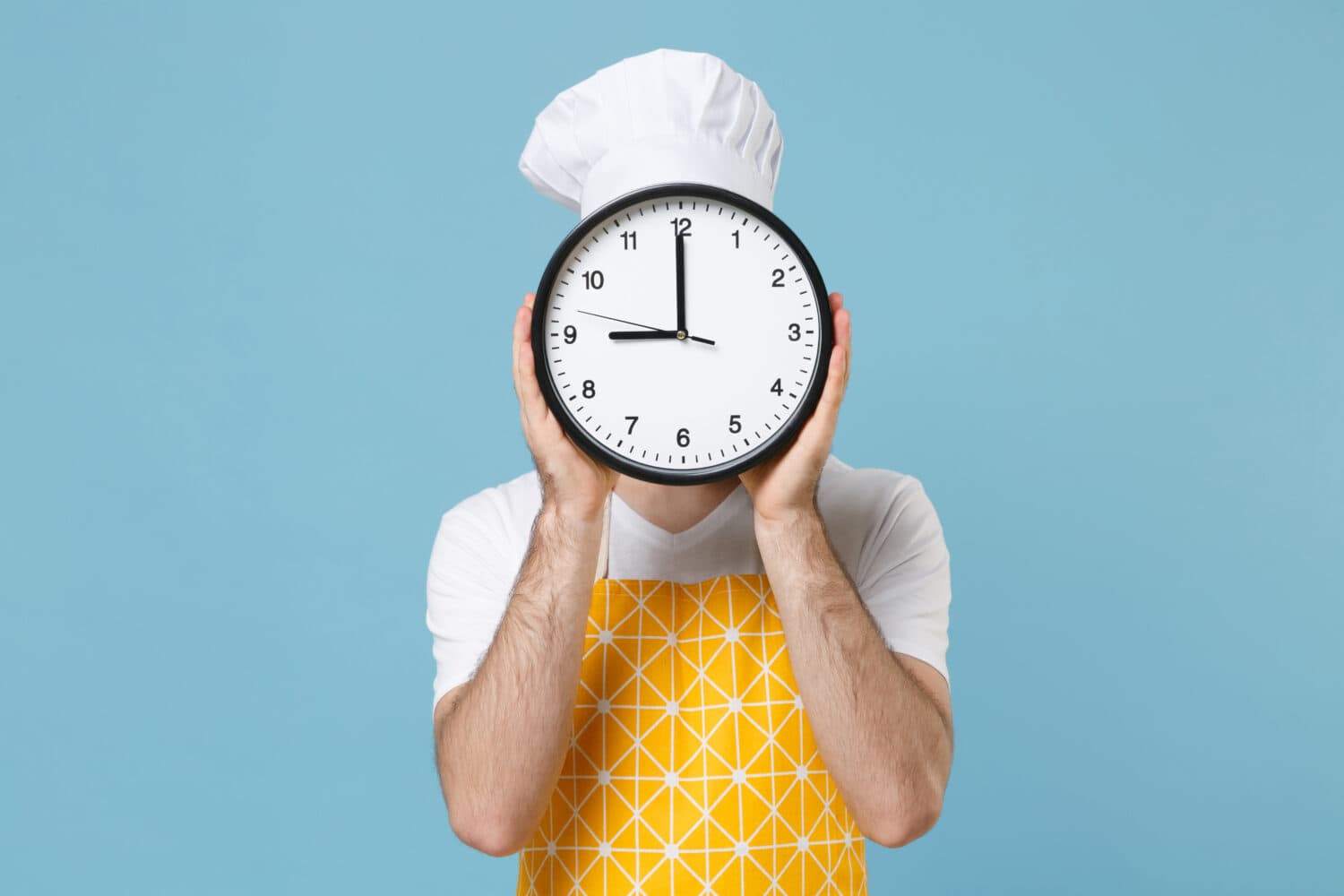 Young bearded male chef or cook baker man in apron white t-shirt toque chefs hat isolated on pastel blue background studio portrait. Cooking food concept. Mock up copy space. Covering face with clock