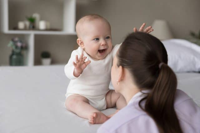 Happy excited baby laughing at mom face, showing positive emotions, waving hands, sitting on bed, playing. New mother talking to charming infant child, cuddling kid, having fun