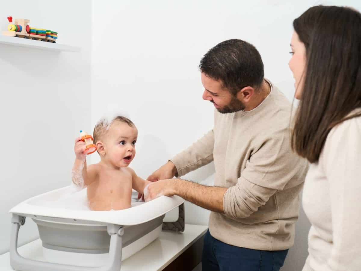 A man and a woman are helping a baby bathe. The baby is in a bathtub and the man is holding a toy