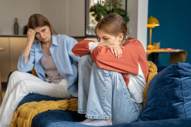 Upset worried mother looking at crying moody teenage daughter, sitting together on sofa, upset frustrated mom and teen girl child not talking after argument, having communication problems