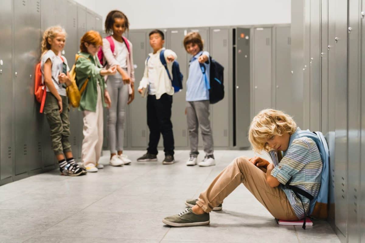 Sad crying schoolboy pupil sitting on the floor at the school hall while his classmates teenagers laughing at him, bullying and torturing him. Social exclusion problem