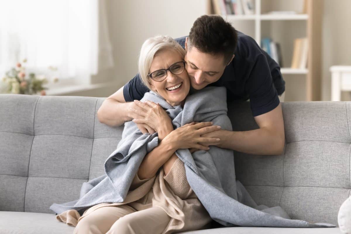 Positive young adult son man caring for happy elderly mature mother sitting on comfortable sofa, throwing warm plaid, scarf on moms shoulders, hugging from behind with love, affection