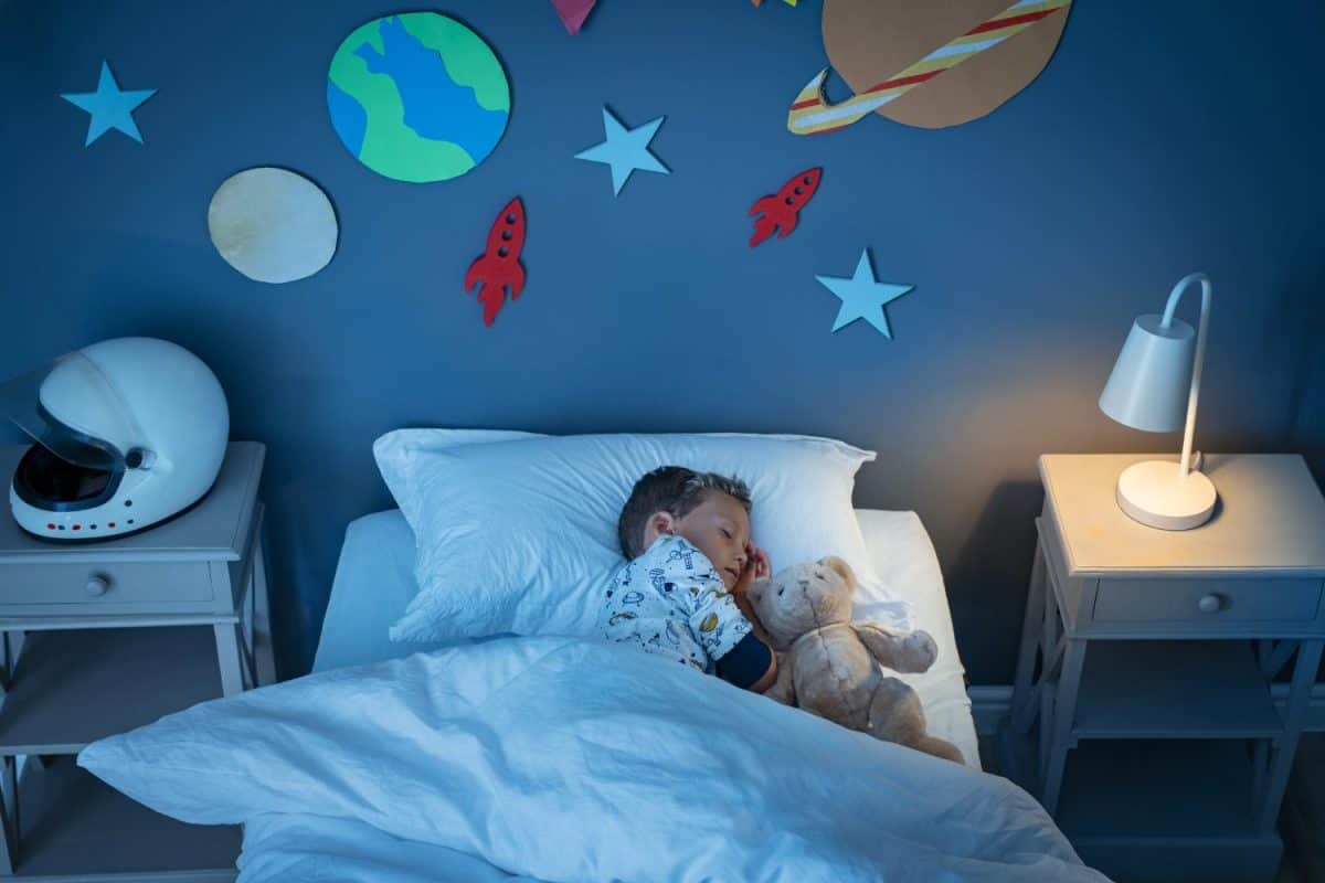 High angle view of little boy dreaming of becoming an astronaut while sleeping with teddy bear in space decorated room. Top view of dreamer child sleeping on bed during the night and the light on.
