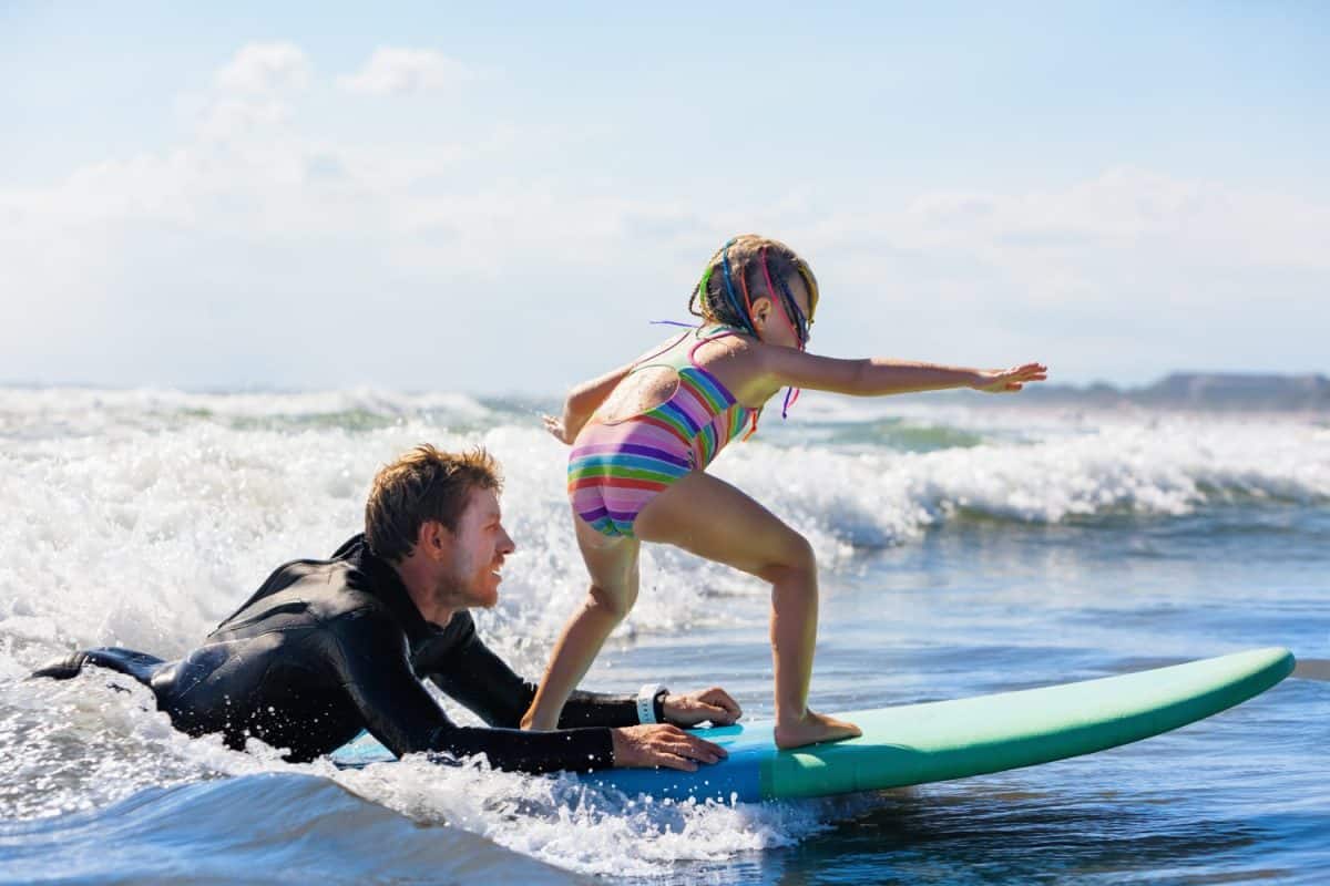 Little surf girl - young surfer learn to ride on surfboard with instructor at surfing school. Active family lifestyle, kids water sport lessons, swimming activity in summer camp. Vacation with child.