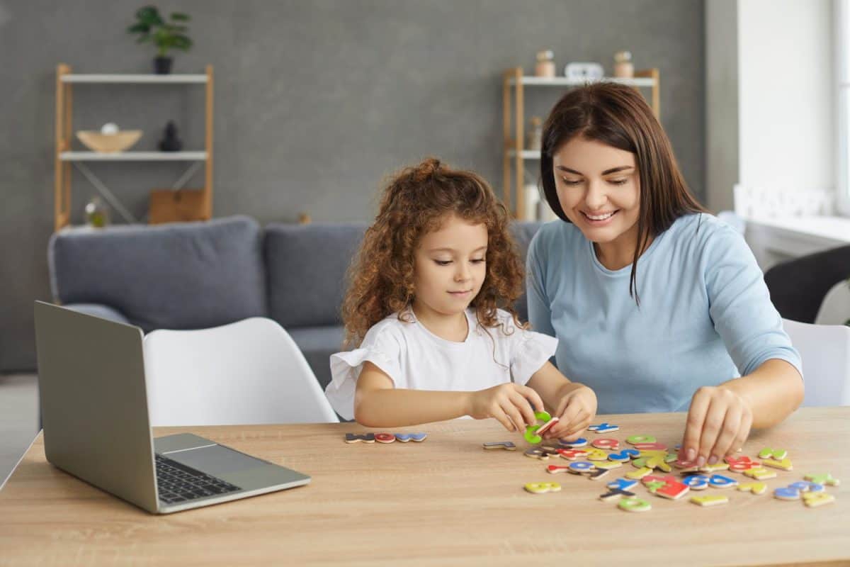 Smiling mother or family tutor helping small child learn foreign language. Happy woman and little girl sitting at desk with laptop and developing English reading skills through fun engaging activities