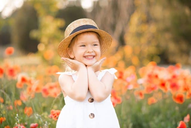 Cute funny baby girl 2-3 year old wear straw hat and white summer dress over flower meadow background. Kid laughing outdoors over nature. Looking at camera. Childhood.