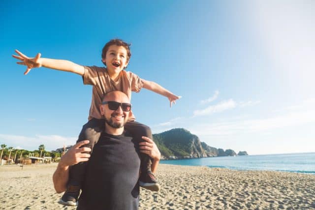 Happy dad and son having fun on beach. Boy sits on his dad's shoulders. Happy childhood and fatherhood. Father-son relationship. Dreams and hope of life at sea. Buying house on coast