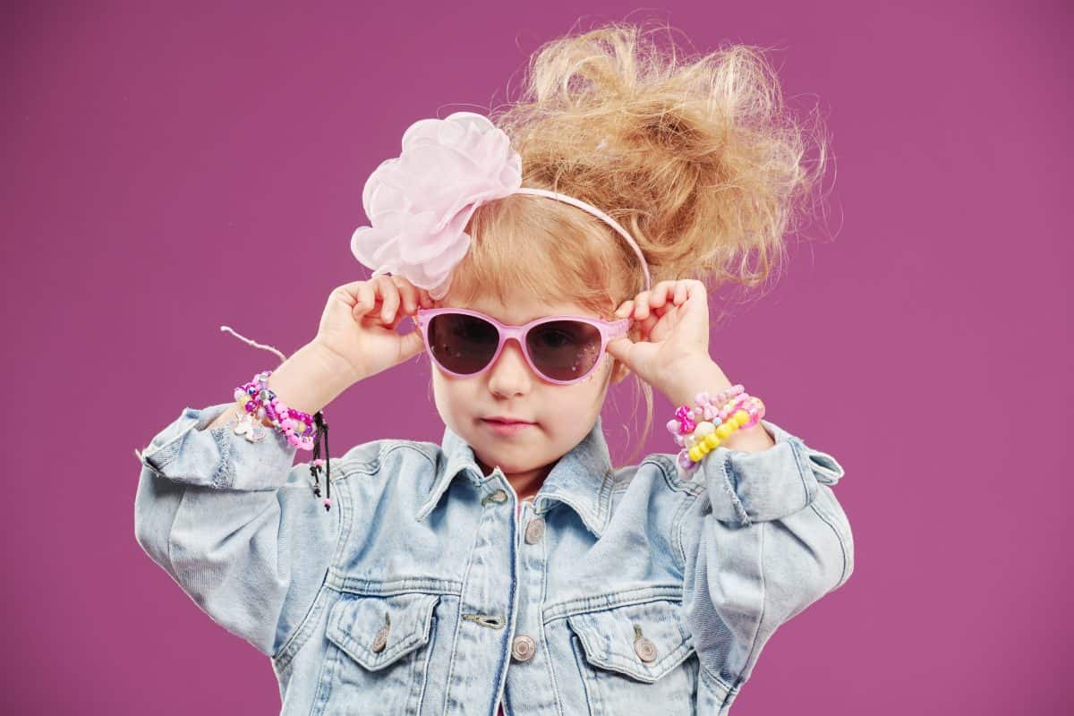 Portrait of a little beautiful cheerful fashionista girl in a denim jacket, pink sunglasses and with a trendy hairstyle. Studio fuchsia background with copy space. Holiday.