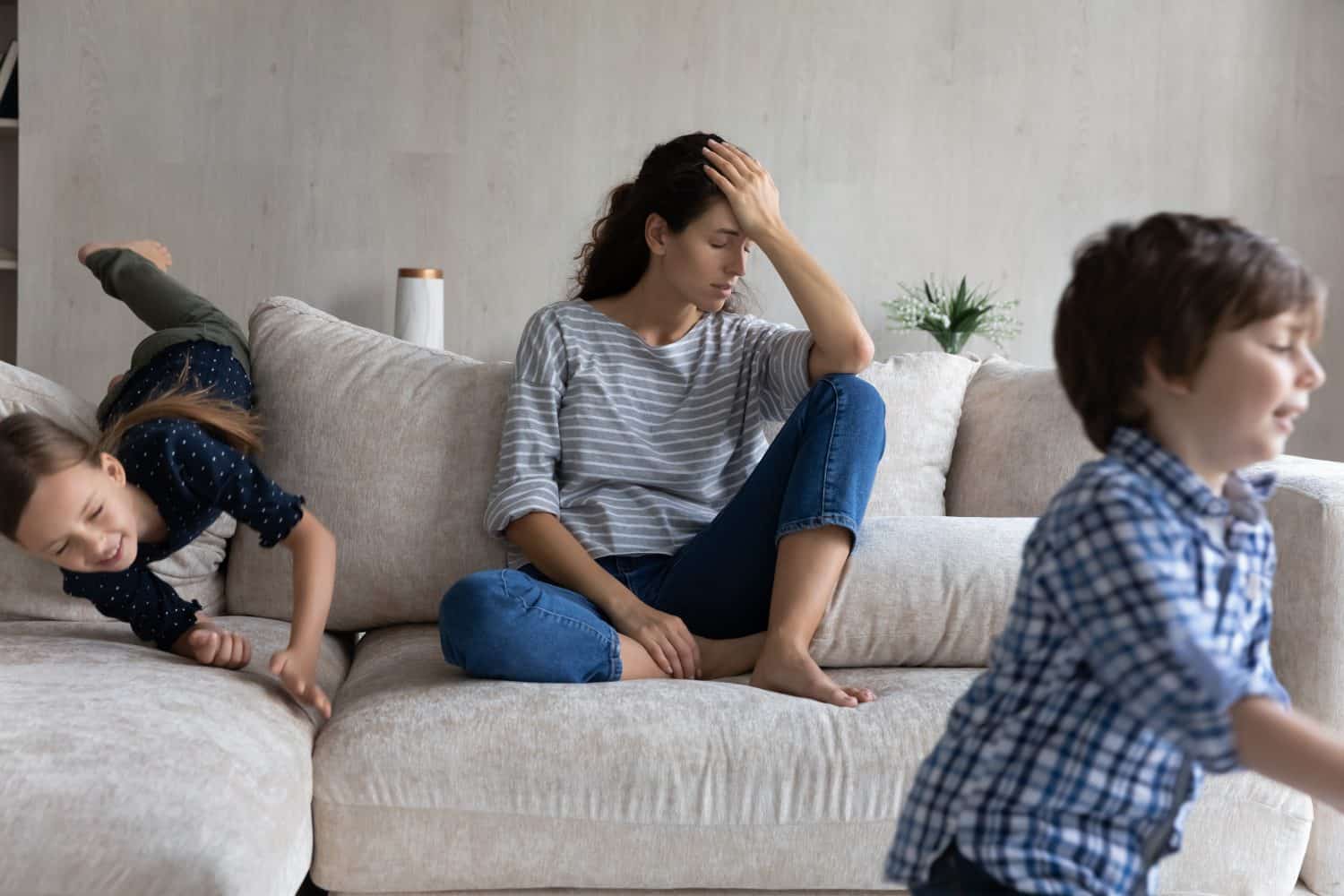 Tired young mother suffering from headache, feeling annoyed and exhausted by noisy kids running around, upset mom or babysitter sitting on couch, having problem with naughty active children