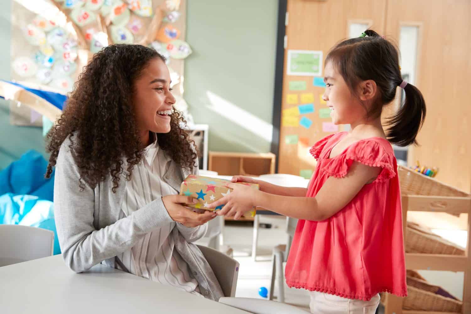 Kindergarten schoolgirl giving a gift to her female teacher in a classroom, side view, close up