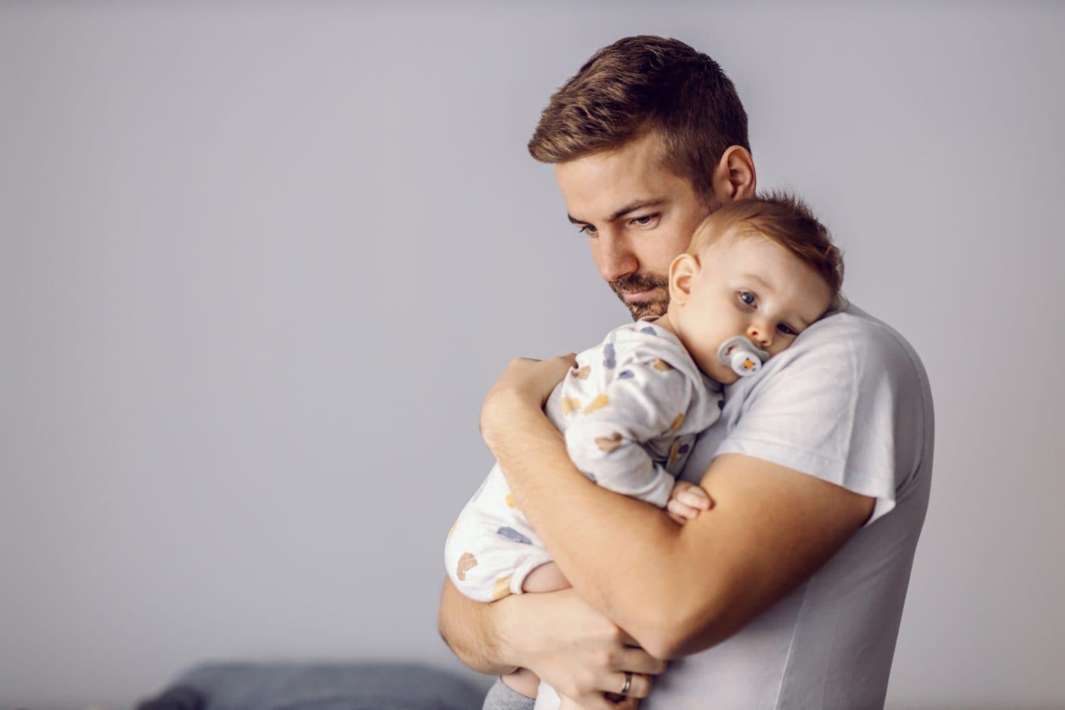 Young good-looking unshaven caring father holding his beloved only son and hugging him. Adorable little boy leaning on father's shoulder, having pacifier in mouth. It's noon nap time