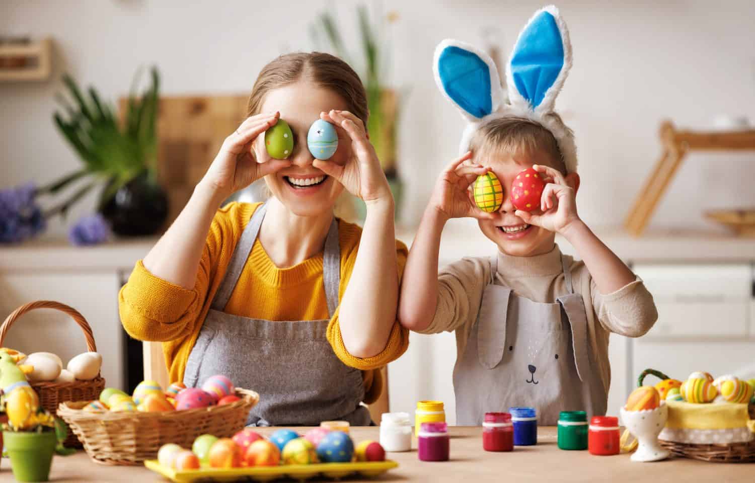 Happy mother and little son wearing aprons holding painted colorful Easter eggs in front of eyes while decorating them with food dyes in cozy kitchen at home. Easter craft activities for families