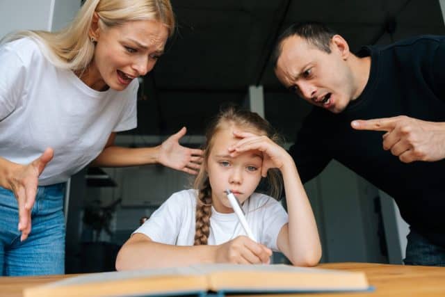 Portrait of angry young parents yelling and scolding together lazy little daughter sitting at table, doing homework, sad looking at camera. Concept of parent disciplining child for bad education.