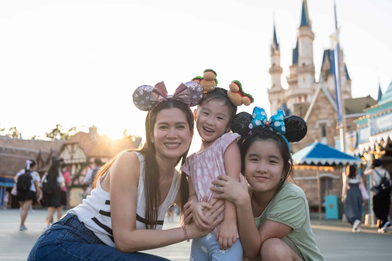 Portrait of Asian mother and two daughter traveling in the theme park. Happy family, attractive woman and children feel happy and relax, spend time for vacation at amusement park and look at camera.