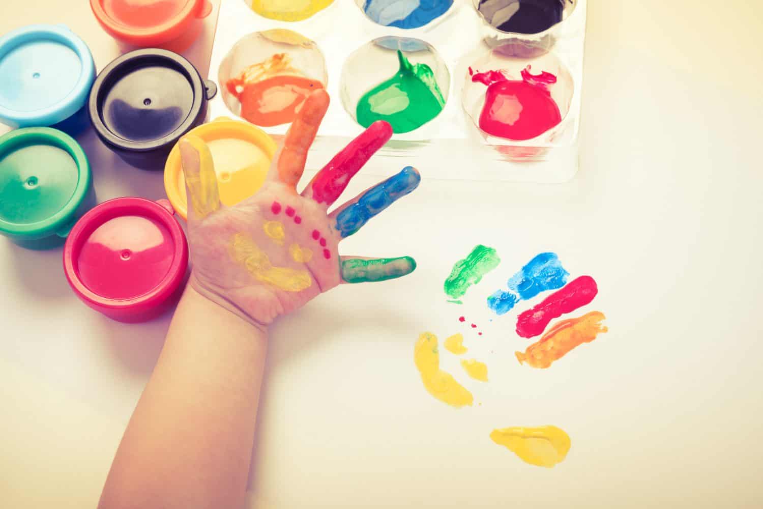 Child paint her palm with smiling face various colors using multicolored drawing tools. Studio shot. Cream tone.