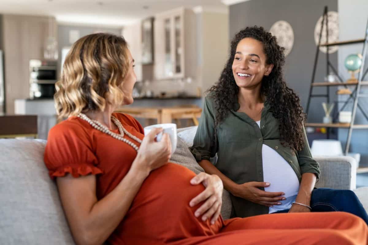 Two multiethnic pregnant women enjoying tea time at home. Smiling pregnant women talking and looking at each other. Happy girls both with a baby bump relaxing on couch and have a friendly conversation