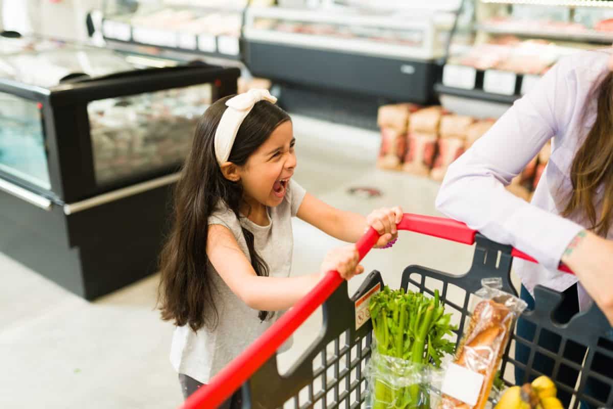 Angry little kid screaming and throwing a tantrum while grocery shopping with her mom at the supermarket because she won't buy her candy