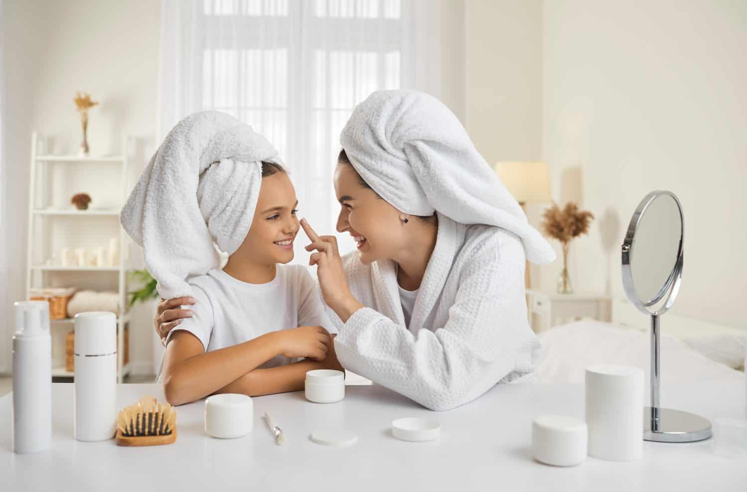 Mother and child having fun during skin care routine. Happy beautiful mommy and pretty little daughter sitting at beauty table, enjoying self care, applying face cream and having good time together