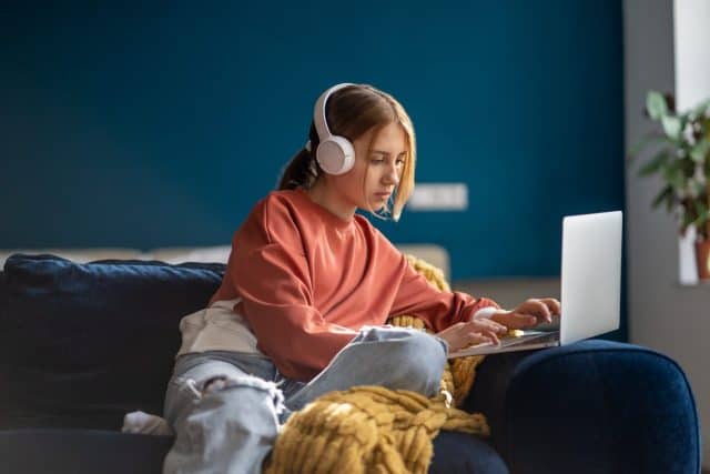 Focused teenage girl wearing headphones typing text on laptop relaxed sitting on couch in living room. Pensive teenager school student studying online, distance learning, using computer at home.