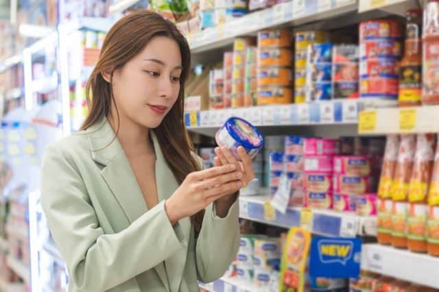 Asian young woman, female in grocery, supermarket shopping food store hand holding tuna can, take out of shelf, read expired date information and ingredients on label, buying healthy products in mall