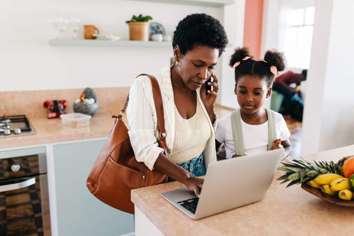 Mother multitasks as she makes a call on her smartphone and uses a laptop while her daughter watches. Working mom going through work tasks before taking her daughter to school in the morning.