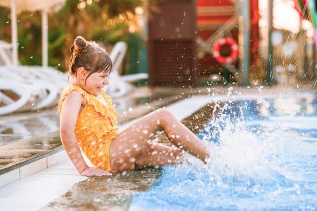 Little preschool girl playing in outdoor swimming pool by sunset. Child learning to swim in outdoor pool, splashing with water, laughing having fun. Family vacations. Healthy children sport activity
