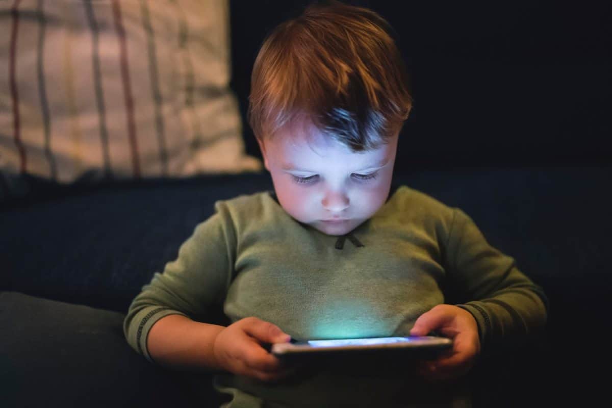 Cute boy sitting on bed watching something on smartphone. Children's screen time.