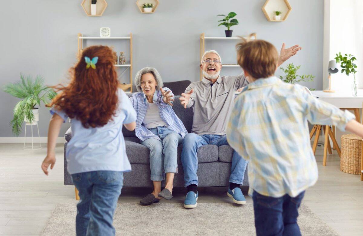 Grandchildren boy and girl running to their senior happy smiling grandparent sitting on sofa at home to hug. Kids enjoying leisure time with grandma and grandpa. Family leisure concept.